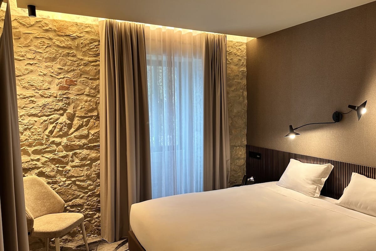 Une chambre d’hôtel avec un grand lit, un mur décoratif en pierre, une fenêtre avec des rideaux légers, un luminaire mural moderne et une chaise.