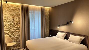 A hotel room with a large bed, a stone accent wall, a window with sheer curtains, a modern wall-mounted light fixture, and a chair.