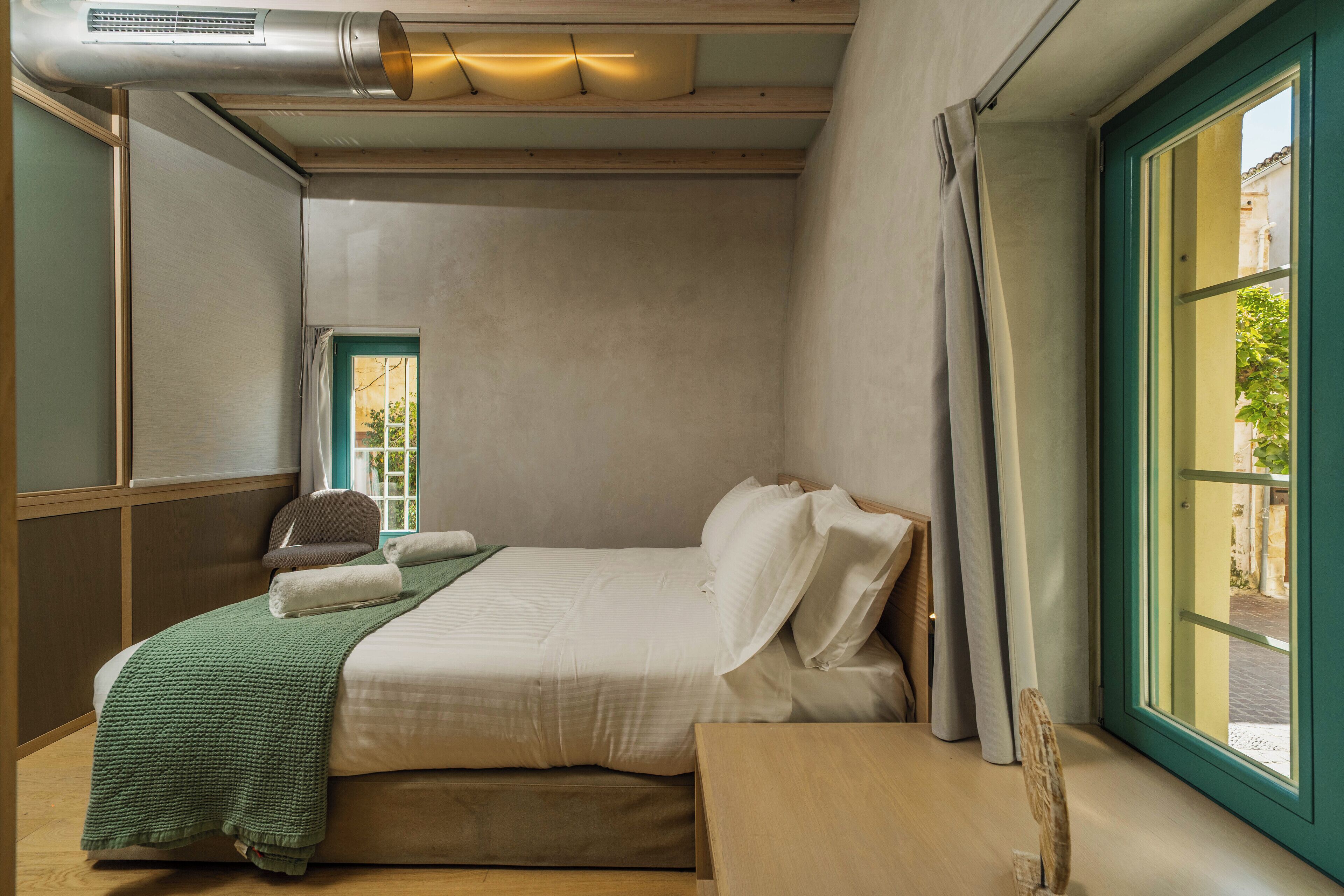 A single bed with a green blanket, a wooden bedside table, and a window with curtains.