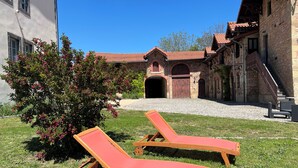 Une cour avec deux fauteuils de jardin en bois, un bâtiment en pierre avec un toit en tuiles et un petit arbre aux fleurs roses.