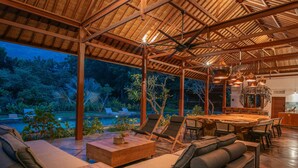A spacious outdoor area with a wooden ceiling, lounge chairs, a dining table, and a view of a pool and greenery. - Experience the Harmony of Luxury, Nature and Wellness! (bali)
