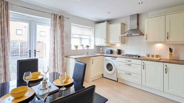 A modern kitchen with a dining area, featuring a dining table set with yellow plates and glasses, a washing machine, and a built-in oven. - Host & Stay | Happy Home (Burton upon Trent)