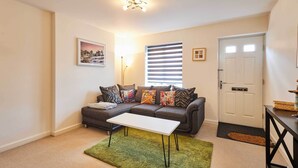 A living room with a sofa, a white coffee table, a green rug, a black console table, and a door with a doormat. - Host & Stay | Happy Home (Burton upon Trent)