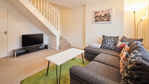A living room with a staircase, a flat-screen TV, a white coffee table, a sofa with patterned cushions, and a framed picture on the wall. - Host & Stay | Happy Home (Burton upon Trent)
