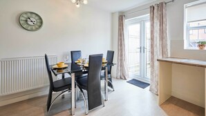 A dining area with a table and chairs, a clock on the wall, and a window with curtains. - Host & Stay | Happy Home (Burton upon Trent)
