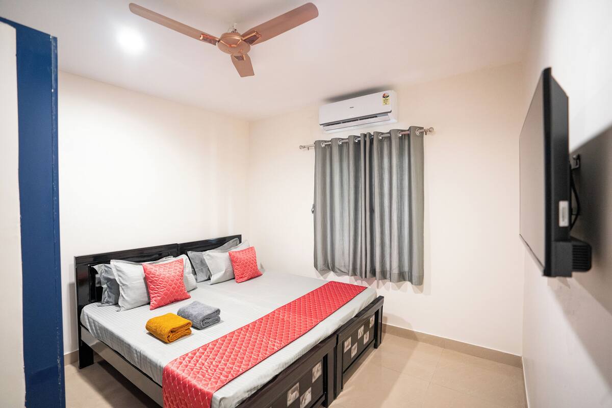 Un dormitorio con cama, ventilador de techo, aire acondicionado, televisor y ventana con cortinas.
