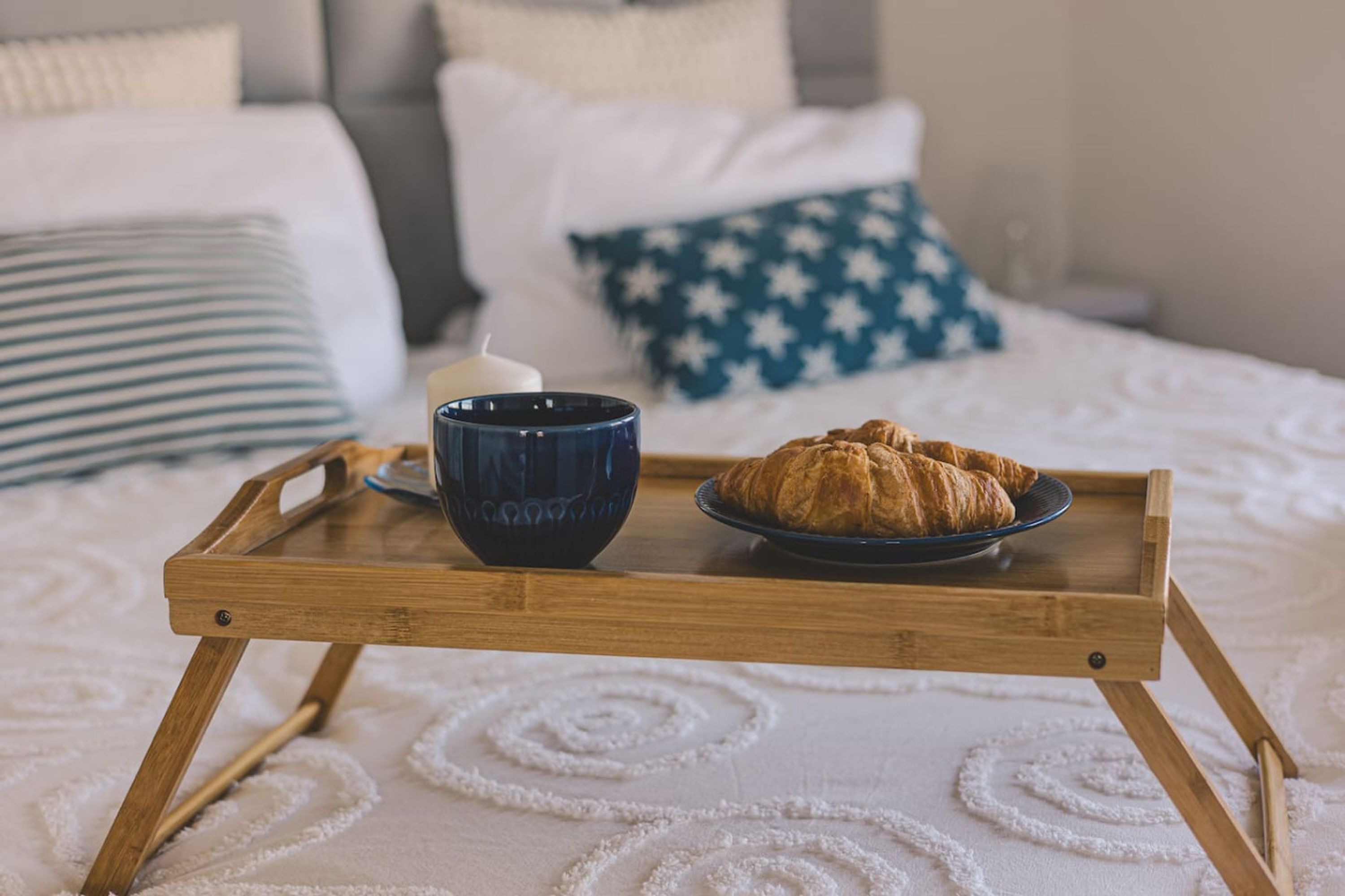 Un plateau en bois avec une tasse bleue et une assiette de croissants, le tout posé sur un lit recouvert d’une couverture blanche et bleue à motifs.