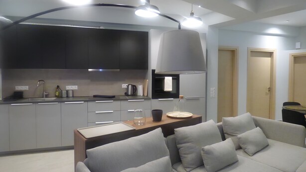 A modern kitchen with grey cabinets, a built-in oven, and a large grey pendant light. There is a wooden dining table with a white placemat and a grey sofa with cushions. - Stylish city Apartment for 4 by MPS (Athina)