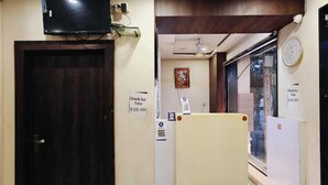 A hotel lobby with a dark wooden door, a flat-screen TV mounted above it, a check-out time sign, a clock, and a green artificial turf rug. - Hotel Jainam Palace (Ujjain)