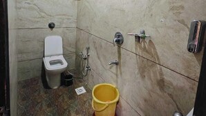A small bathroom with a toilet, a shower, and a yellow bucket on the floor. - Hotel Jainam Palace (Ujjain)