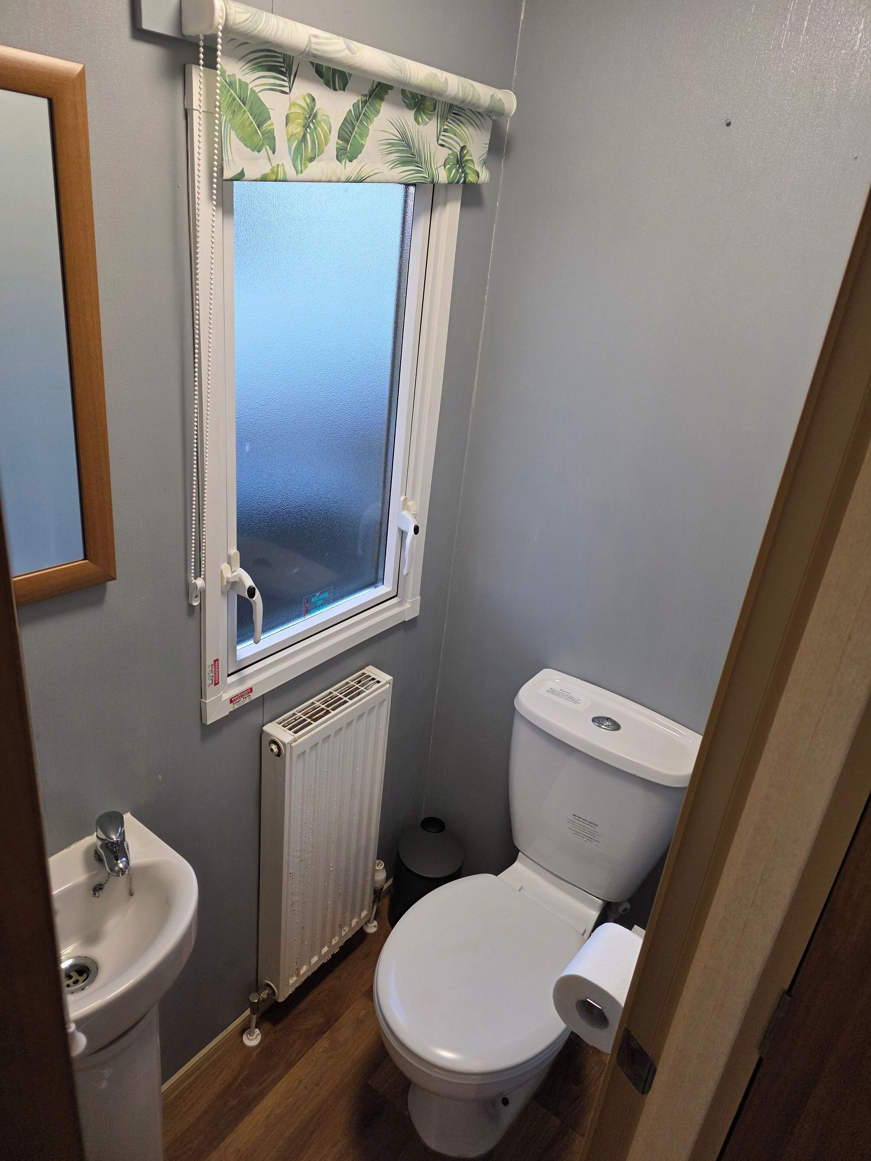 Une petite salle de bain comprenant des toilettes, un lavabo, une fenêtre avec un rideau à motifs de feuilles et un radiateur.