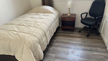 Un lit simple avec une couette beige, une petite table de chevet en bois avec une lampe, une chaise de bureau noire et une fenêtre avec des rideaux.