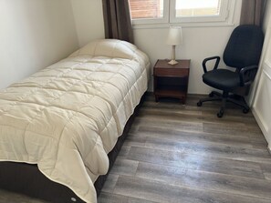 Een eenpersoonsbed met een beige dekbedovertrek, een klein houten nachtkastje met een lamp, een zwarte bureaustoel en een raam met gordijnen.