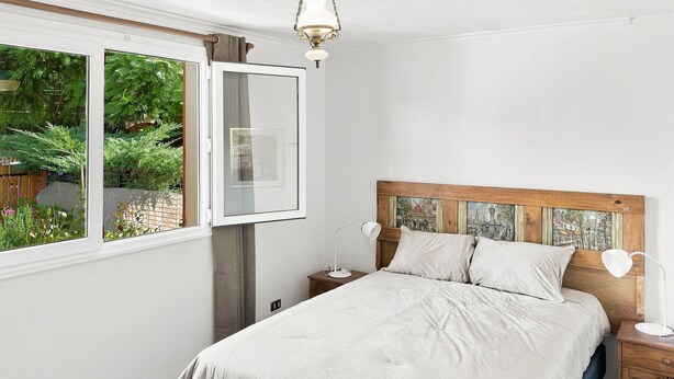 A bedroom with a bed, bedside tables, a window with a view of greenery, and a headboard with a cityscape painting. - Oceana Suites Casa Ñuñoa (Santiago)