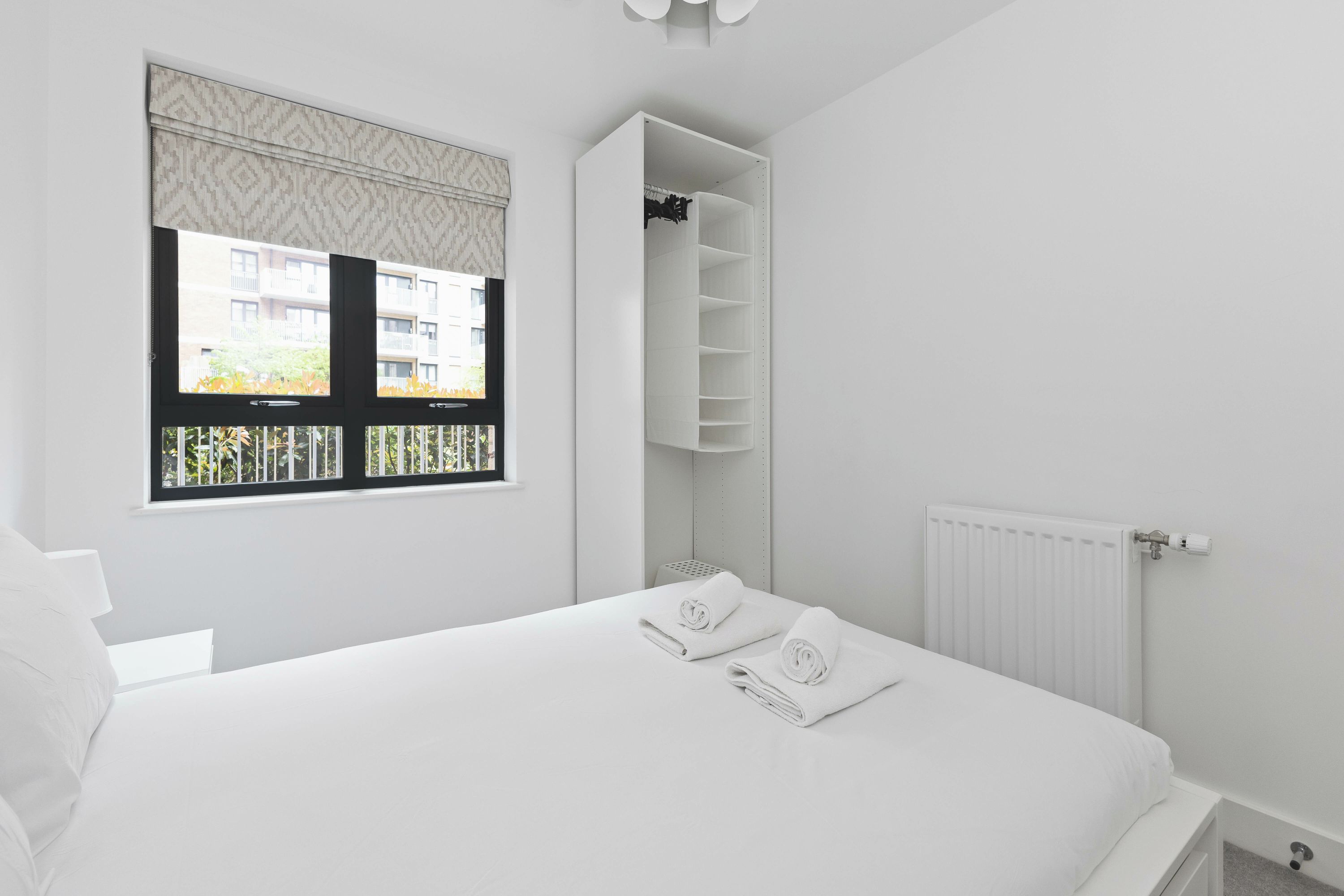 A bedroom with a bed, a window with blinds, a radiator, and a built-in wardrobe.