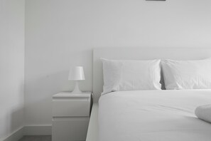 A minimalist bedroom with a single white bedside table, a white lamp, and a neatly made bed with white bedding. - Modern 2 Bed 2 Bath with Patio | Close to Wembley (Greater London)