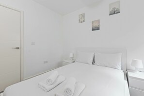 A white hotel room with a bed, two bedside tables, a nightstand, and a door.
