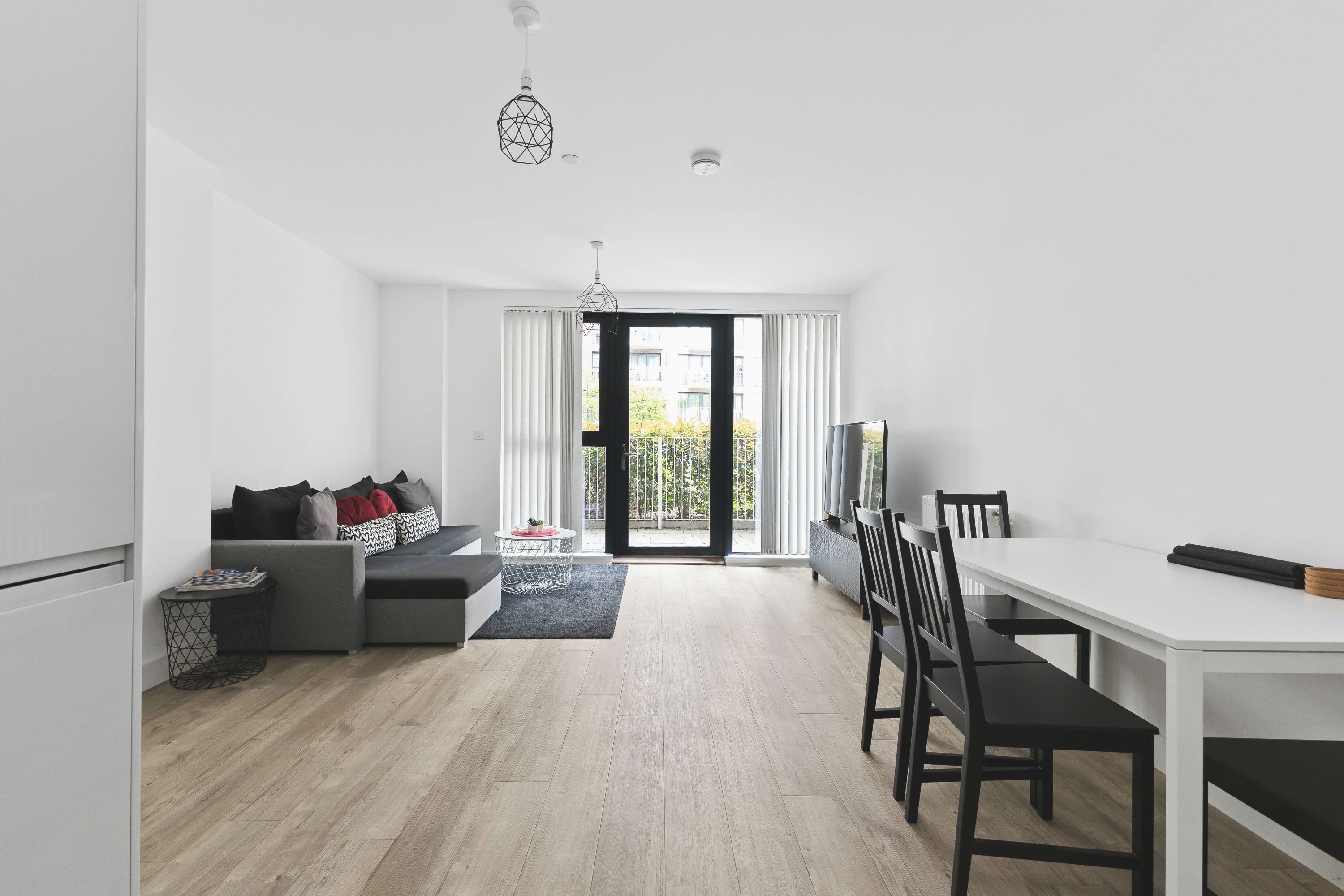 A modern living room with a sofa, a dining table, and chairs. There is a large window with curtains, a ceiling light, and a view of the outside.