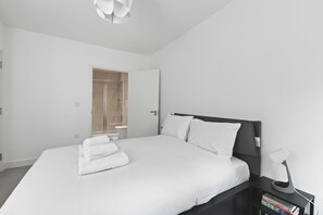 A modern bedroom with a large bed, a bedside table with a lamp, and a shower area visible through an open door. - Modern 2 Bed 2 Bath with Patio | Close to Wembley (Greater London)