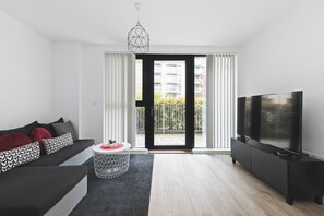A modern living room with a sofa, a TV, and a view of a building through the glass door. - Modern 2 Bed 2 Bath with Patio | Close to Wembley (Greater London)
