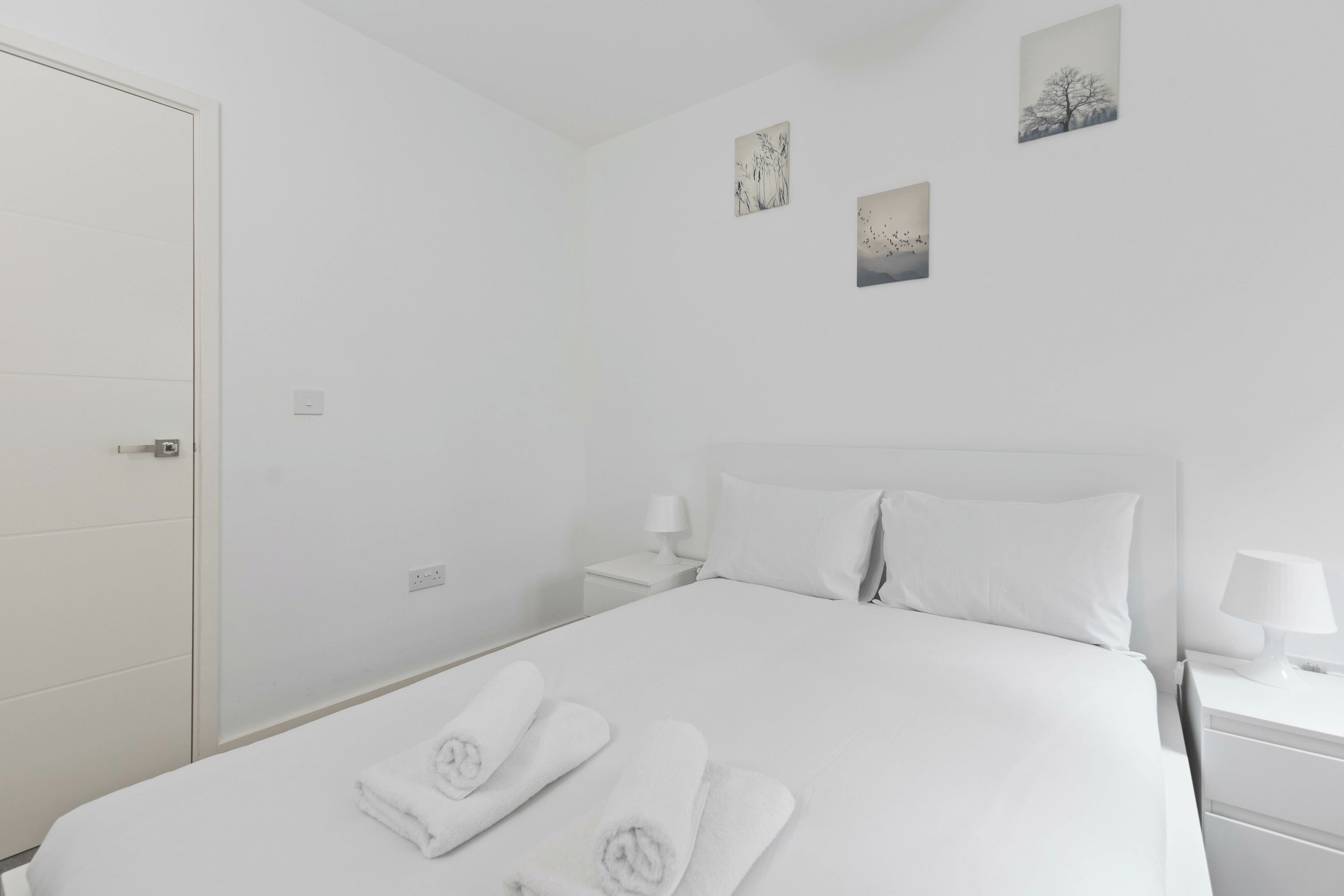 A white hotel room with a bed, two bedside tables, a nightstand, and a door.