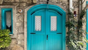 Μια διπλή πόρτα σε τιρκουάζ χρώμα, διακοσμημένη με δαντελένια διακοσμητικά στοιχεία, είναι ενσωματωμένη σε ένα πέτρινο κτίριο, το οποίο διαθέτει ένα μικρό παράθυρο και ένα κουδούνι.