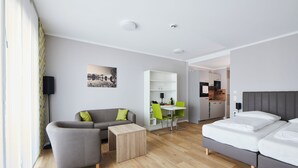 A modern hotel room with a grey sofa, a small wooden table, a bed with white linens, and a dining area with green chairs. - ADAPT APARTMENT HOTEL Wetzlar (Wetzlar)