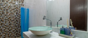 Casa de banho com lavatório de vidro, duas torneiras, cortina de chuveiro azul e produtos de higiene num tabuleiro.