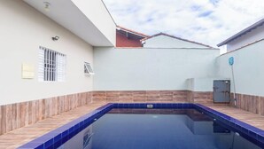 A swimming pool with tiled edges, a wooden deck, and a white building with a tiled roof. - Casa em Peruíbe a 250m da Praia com Piscina (Peruíbe)