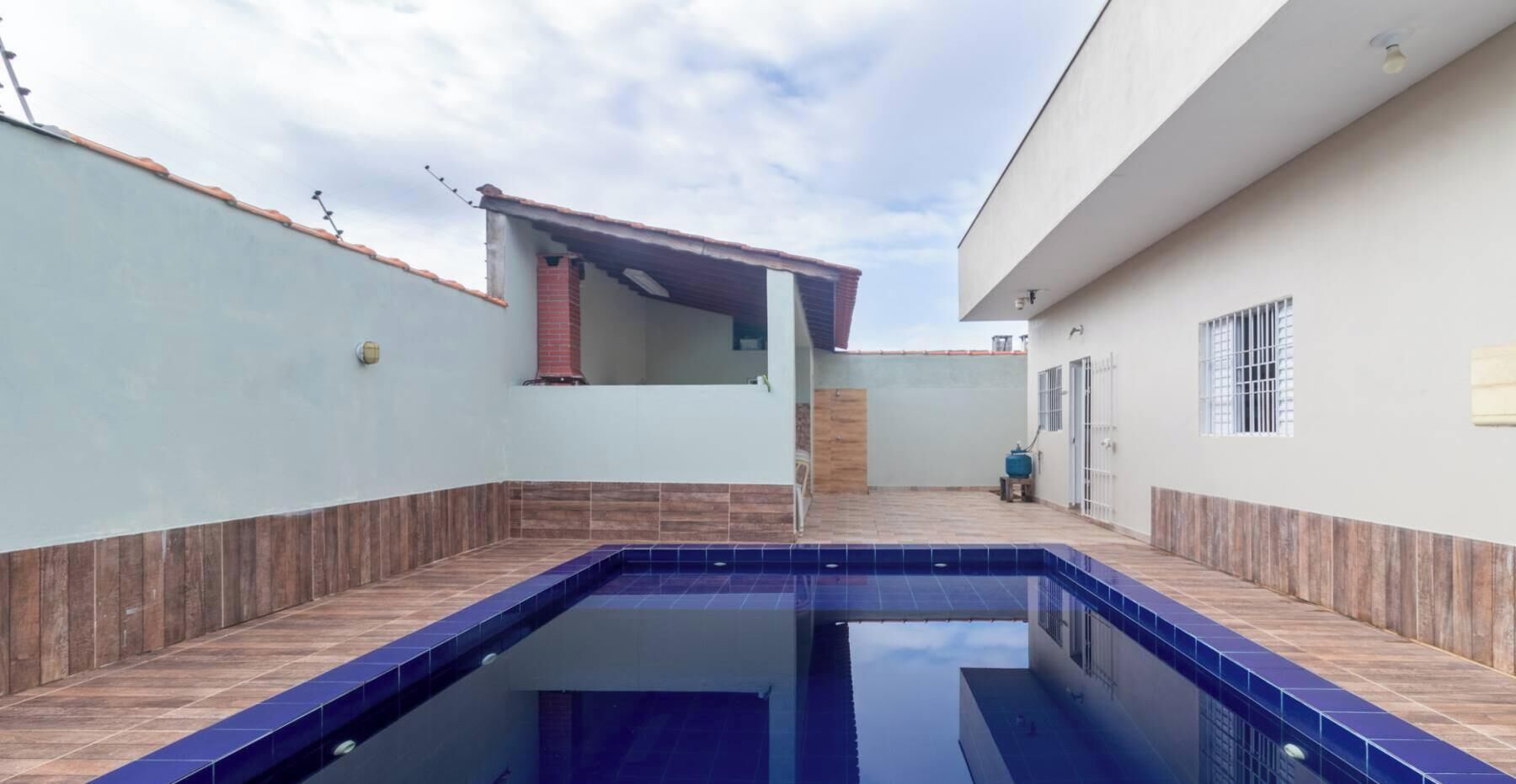 Une piscine avec des bords carrelés, une cheminée en briques et un bâtiment blanc doté d’une fenêtre et d’une porte.