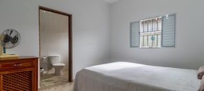 Quarto com cama, cómoda de madeira, ventoinha, sanita e janela com grades.