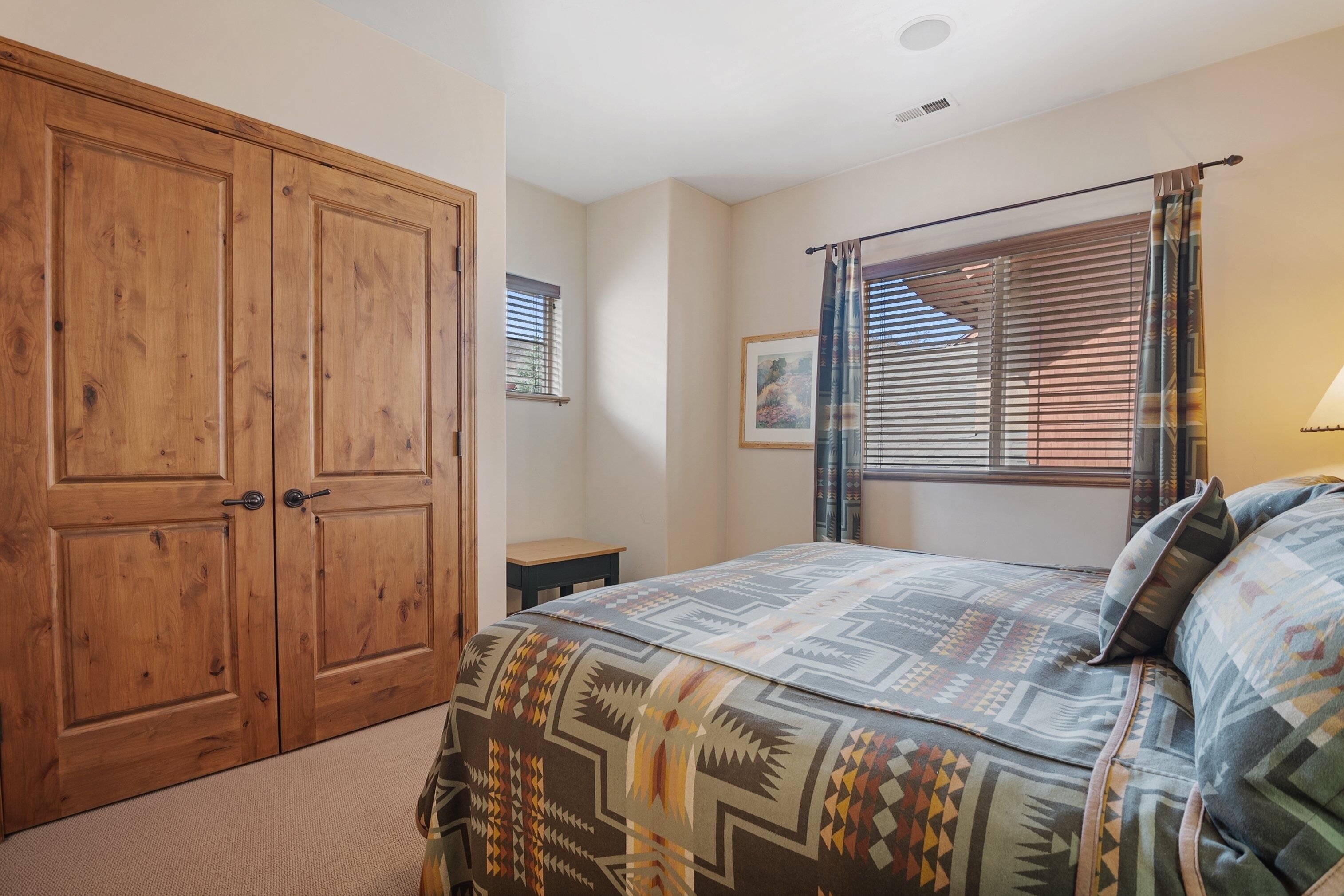 A bedroom with a large wooden wardrobe, a bed with patterned bedding, a bedside table, and a window with blinds.