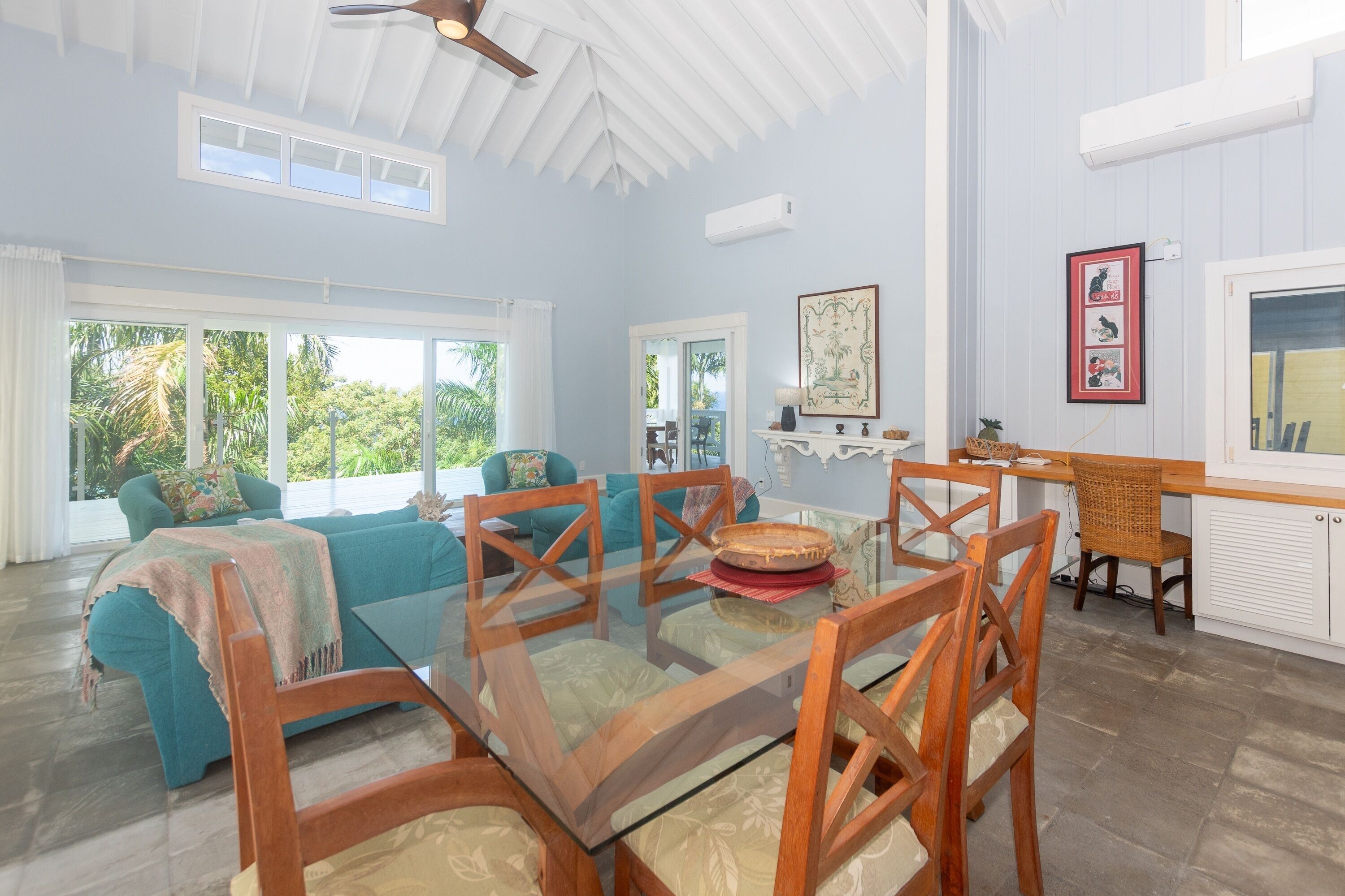 A spacious living area with a glass dining table, teal sofas, and a kitchen area with a white countertop and hanging artwork.