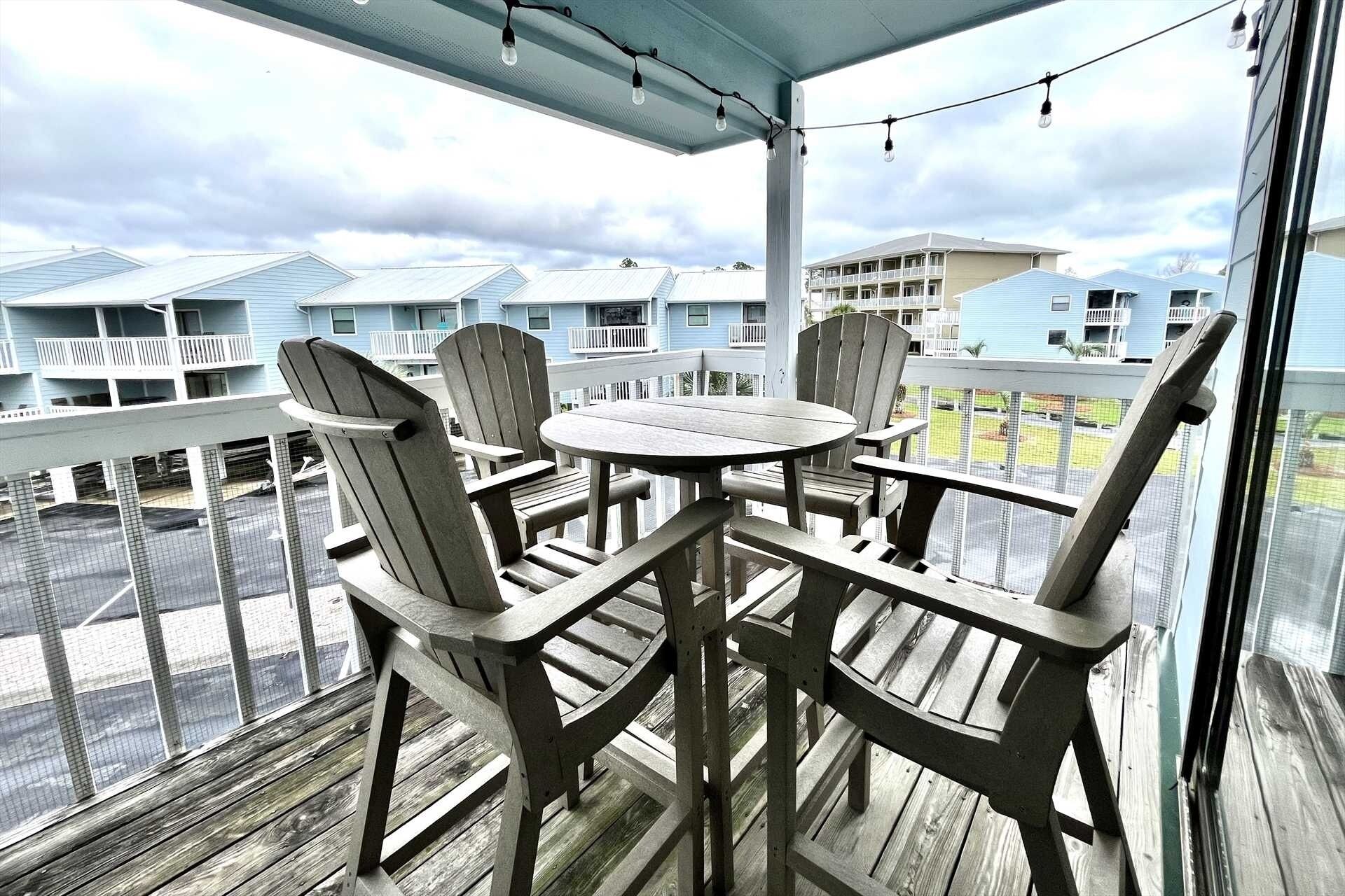 A covered outdoor seating area with Adirondack chairs and a round table, overlooking a row of blue buildings.