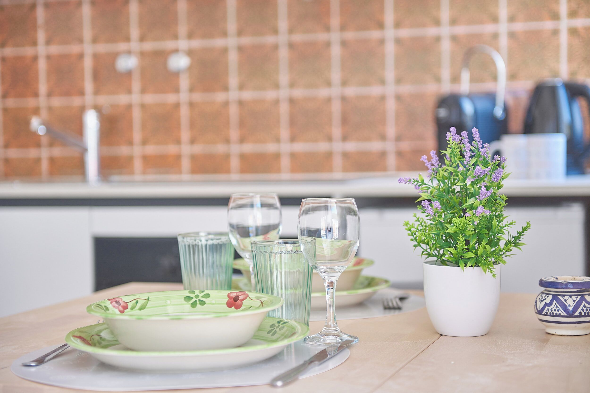 Une cuisine avec un mur recouvert de carreaux, une plante en pot et un ensemble de vaisselle comprenant des assiettes, des verres et des couverts.