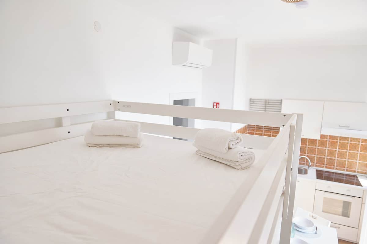 Une chambre blanche avec un lit simple, donnant sur une cuisine et une salle de bain.