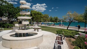 A fountain with multiple tiers, surrounded by benches and greenery, with a view of a body of water and distant buildings. - Suite in the heart of the city, just a few meters from the sea (Kaštel Stari)