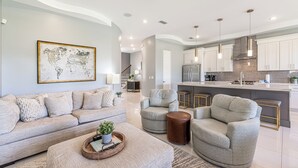Una sala de estar moderna con un sofá, sillones y una mesa de centro. Se ve una cocina con armarios blancos y una isla grande al fondo.