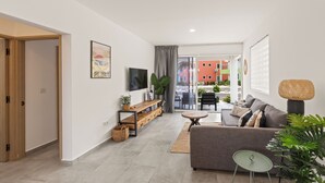 Una sala de estar moderna con un sofá gris, una mesita redonda pequeña y un televisor en la pared. Se ve un mueble de madera con una planta y un marco de fotos. Al fondo, una ventana grande con cortinas.