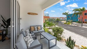 Un área de estar moderna al aire libre con un sofá y una mesa, un letrero que dice "Beach Life", y vista a una calle con palmeras y edificios coloridos.