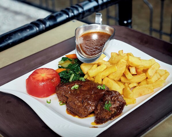 A plate of food including steak, fries, a tomato, and greens, served on a white plate with a side of sauce. - Acacia country Inn (Mbarara)