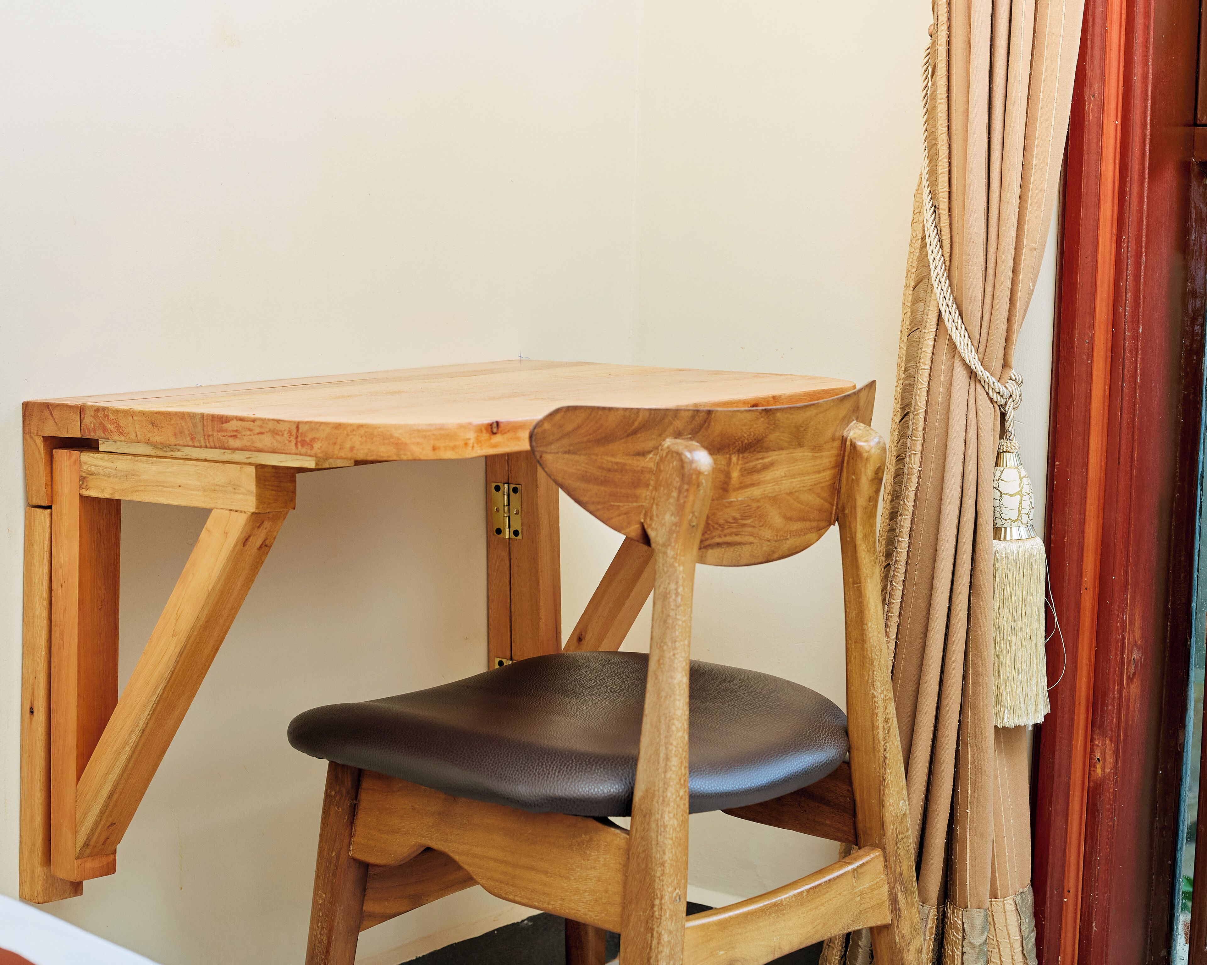 A wooden table and chair with a simple design, positioned against a plain wall with a curtain partially visible.