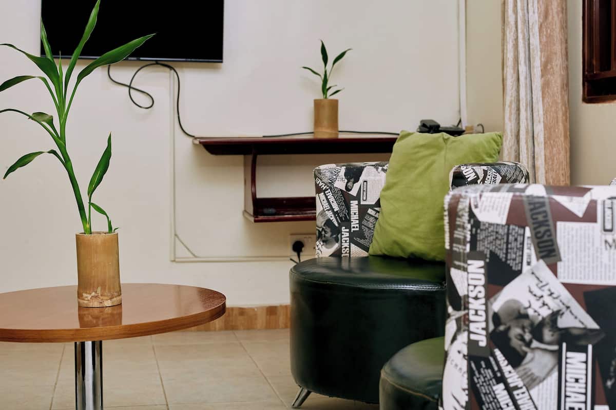 A living room with a round wooden table, a green sofa with patterned cushions, a black armchair, and a potted plant.