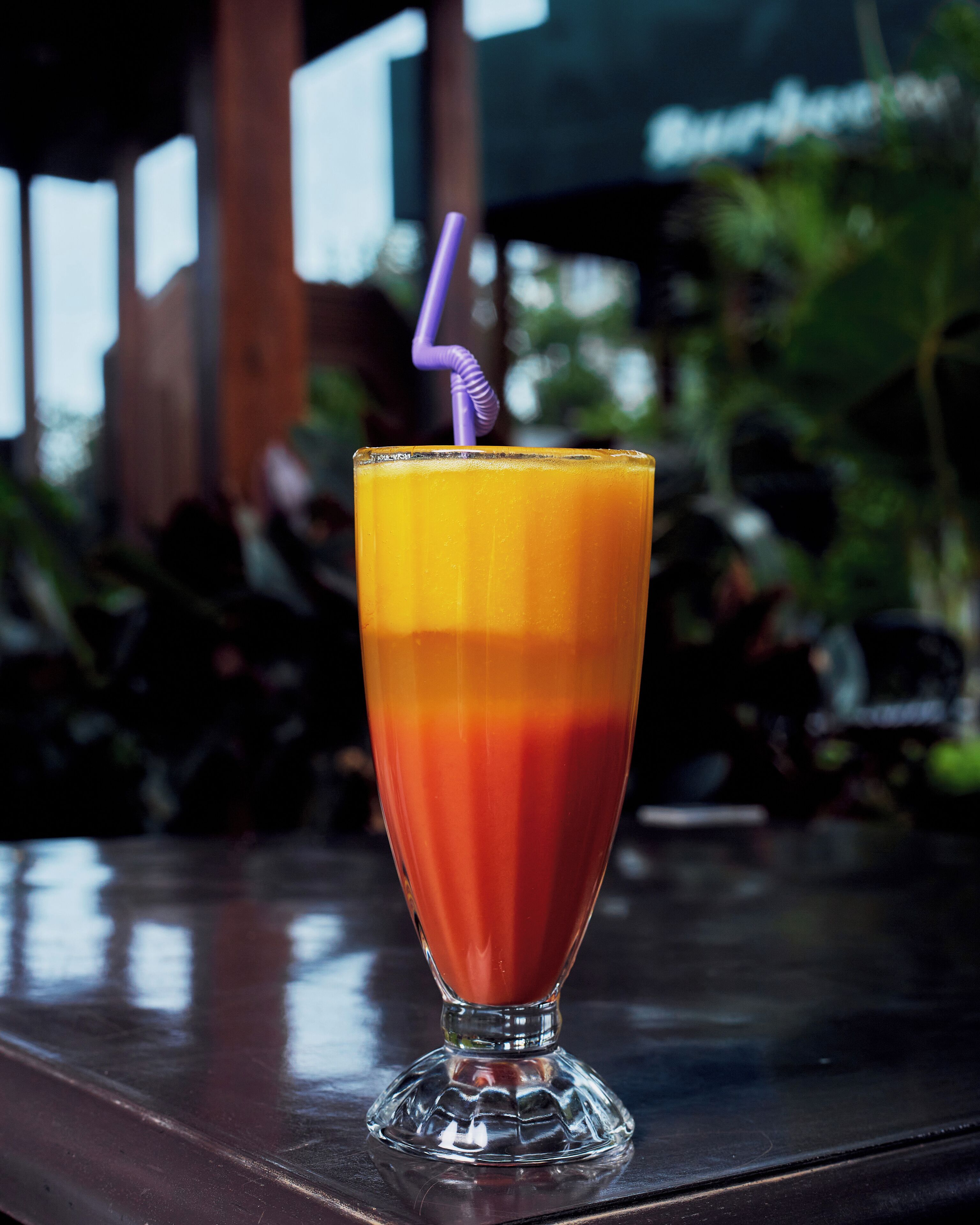 Ein Glas Orangensaft mit einem violetten Trinkhalm auf einer reflektierenden Oberfläche.