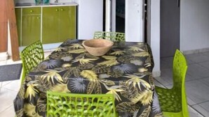 Un coin repas avec une table recouverte d’un motif de feuilles tropicales, entourée de chaises vertes. On aperçoit au fond un plafond en bois et un meuble vert contenant un four à micro-ondes.