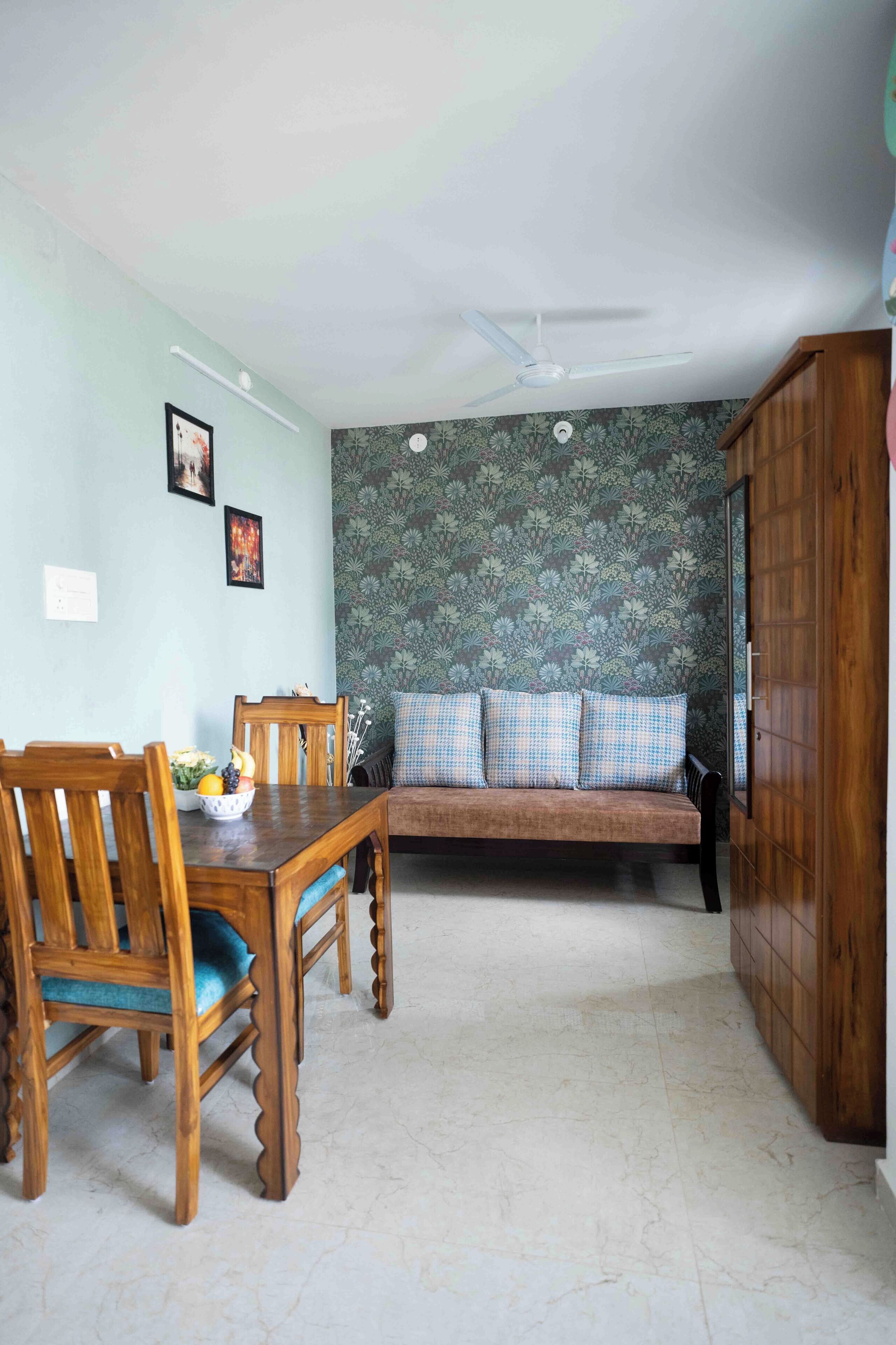 A dining area with a wooden table and chairs, a sofa with checkered cushions, and a patterned wallpaper wall.