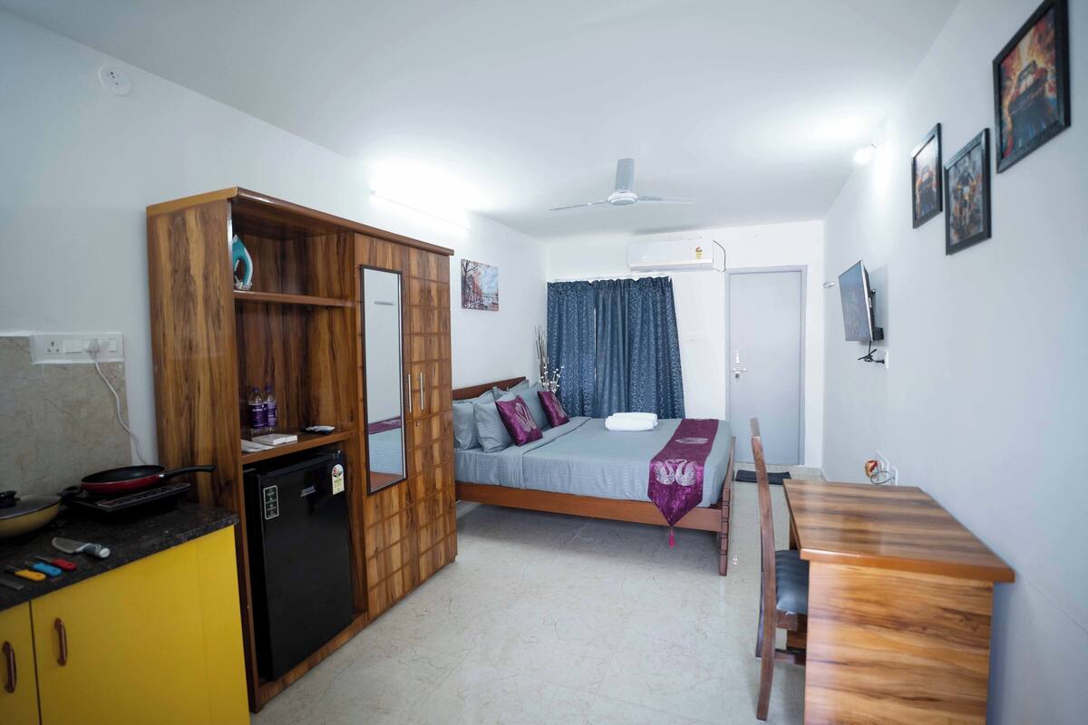 Una habitación compacta con cama, un armario de madera, una pequeña cocina con refrigerador y una mesa de comedor.