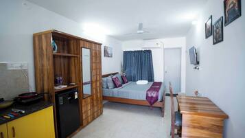 A compact bedroom with a bed, a wooden wardrobe, a small kitchen area with a refrigerator, and a dining table.