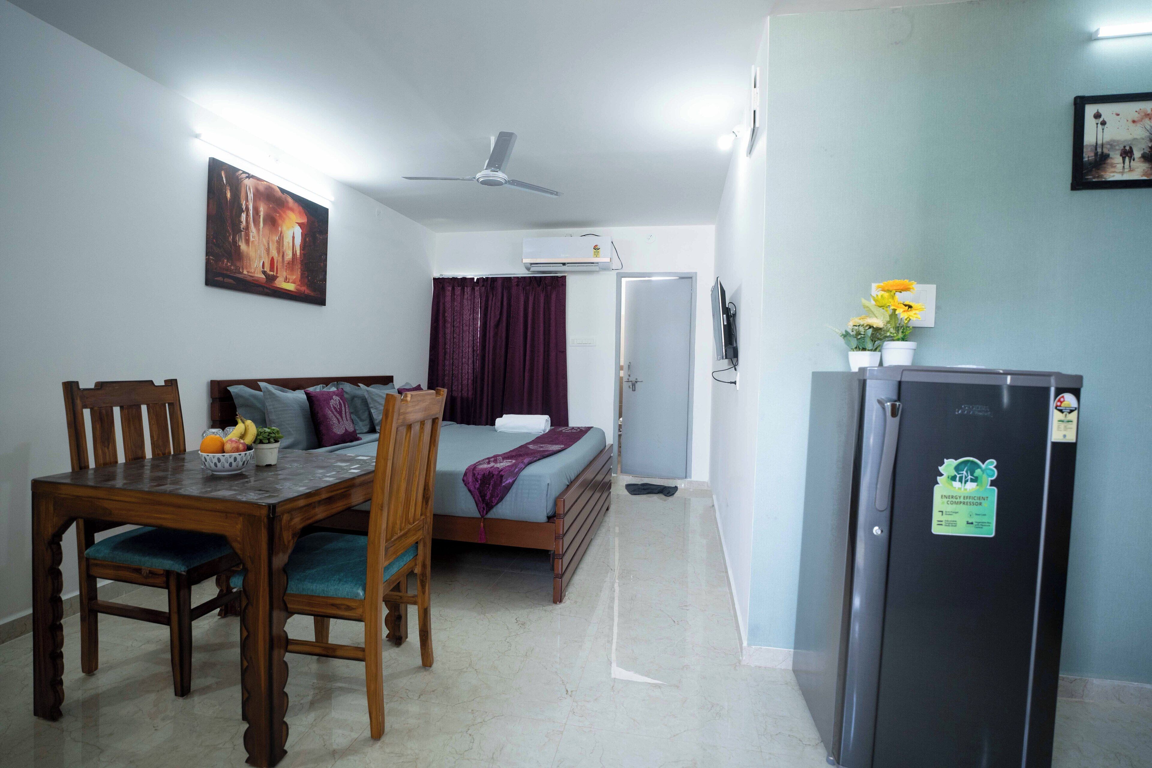 A compact hotel room with a bed, a dining table with chairs, a refrigerator, a ceiling fan, and a painting on the wall.