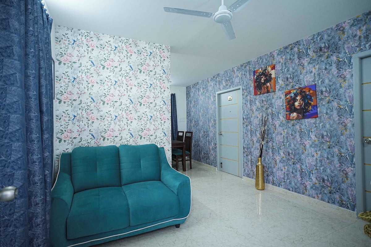 Una habitación con un sofá azul verdoso, papel tapiz floral, un ventilador de techo y dos cuadros en la pared.